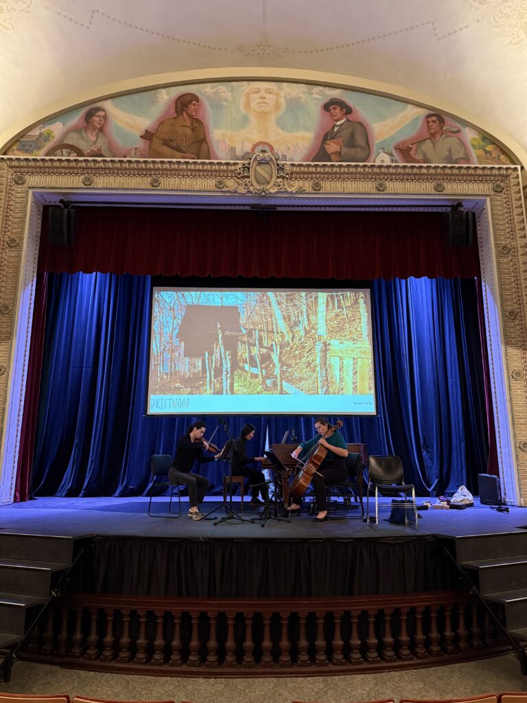 Violinist Heather Thomas, cellist Cecilia Huerta-Lauf, and pianist Sebastian Chang on stage at The Carnegie in Convington Kentucky, performing under a proscenium arch created by Harlan Hubbard called The Spirit of Convington