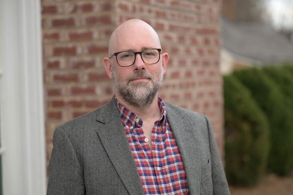 A man with glasses and a beard wearing a gray blazer, red shirt standing in front of a brick wall